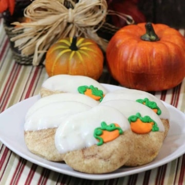 Pumpkin cookies white white icing and a pumpkin design on a white plate with pumpkins in background.