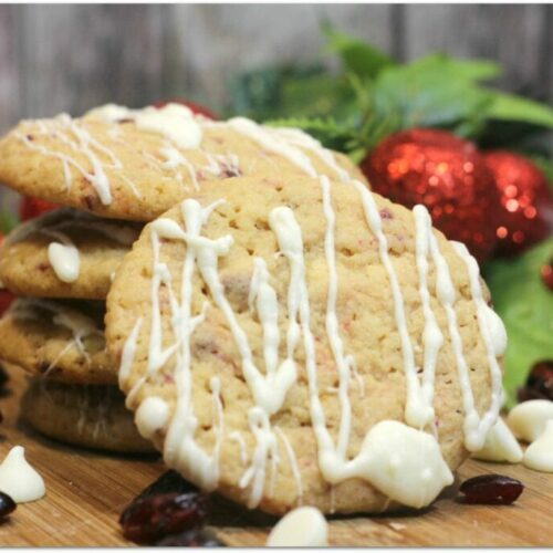 Cookies studded with cranberries and drizzled with white chocolate on a wood board with Christmas decor in the background.