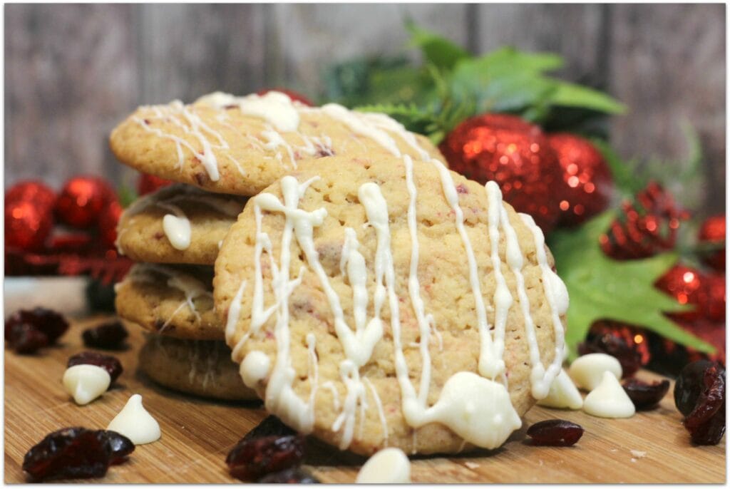 Cookies studded with cranberries and drizzled with white chocolate on a wood board with Christmas decor in the background.