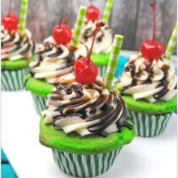 green cupcakes with vanilla frosting, chocolate syrup, cherry, and a straw