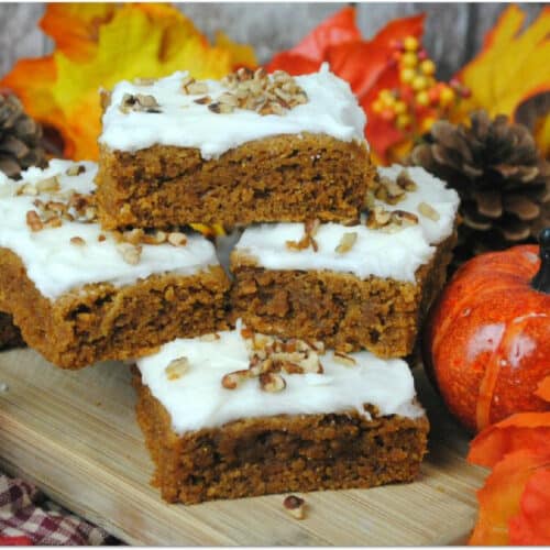Pumpkin bars with cream cheese frosting on a wood board with fall leaves and pumpkin.