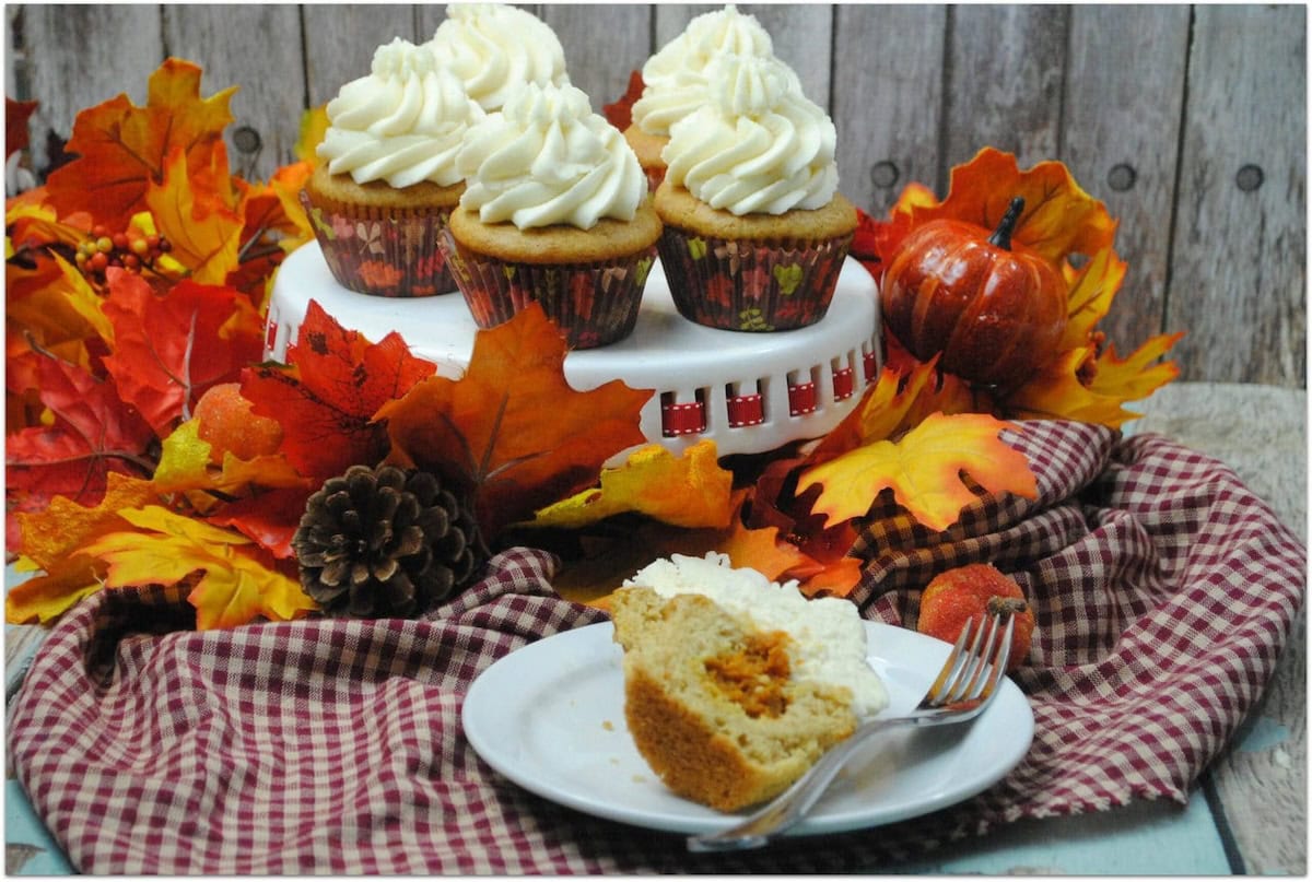 Split pumpkin pie cupcake with cream cheese frosting on a plate with more cupcakes on a cake stand in back with fall leaves.