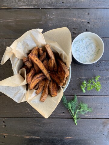 crispy oven sweet potato fries in a paper lined basket, with dip and herbs on a black board
