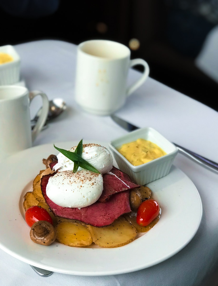 Eggs, ham, potatoes, and tomatoes on a white plate with coffee cup in background.