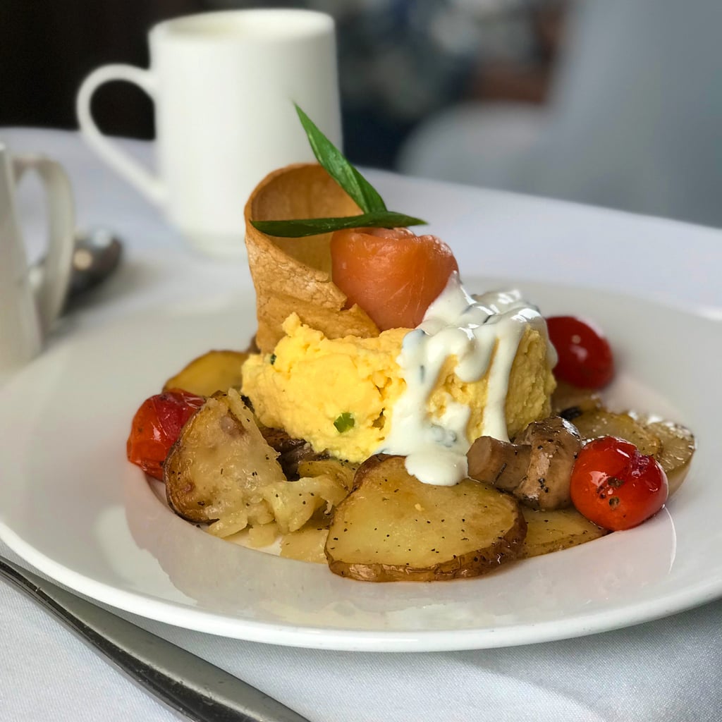 Eggs, potatoes, tomatoes, and creme fraiche on white plate on white tablecloth with coffee cup in background