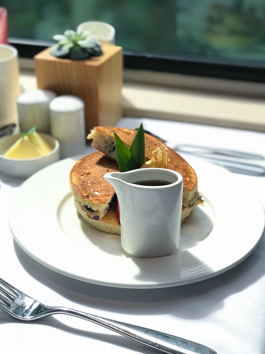 Pancakes with cup of syrup on white plate in front of window.