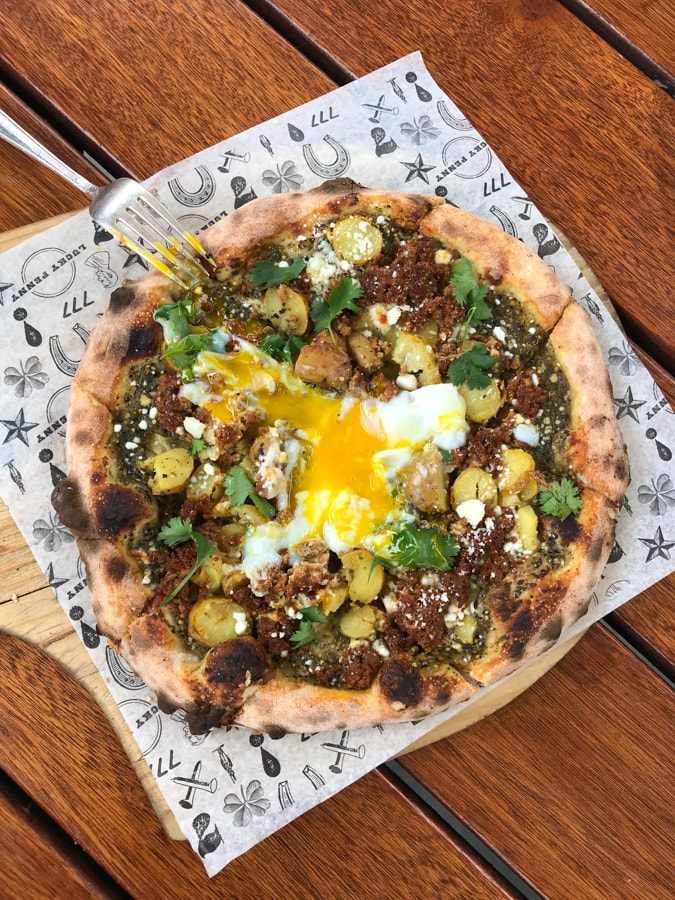 Pizza with meat and potatoes and egg on top on paper on wood table.