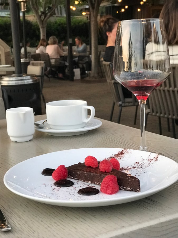 Chocolate torte with raspberries on a white plate with a glass of red wine and coffee.