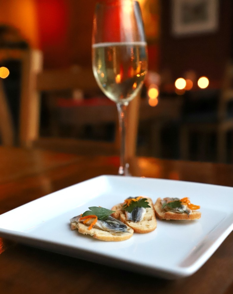 Fish on crostini on white plate with glass of wine in background.