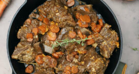 Guinness braised short ribs with fresh thyme in a cast iron skillet with carrots and onion in background.