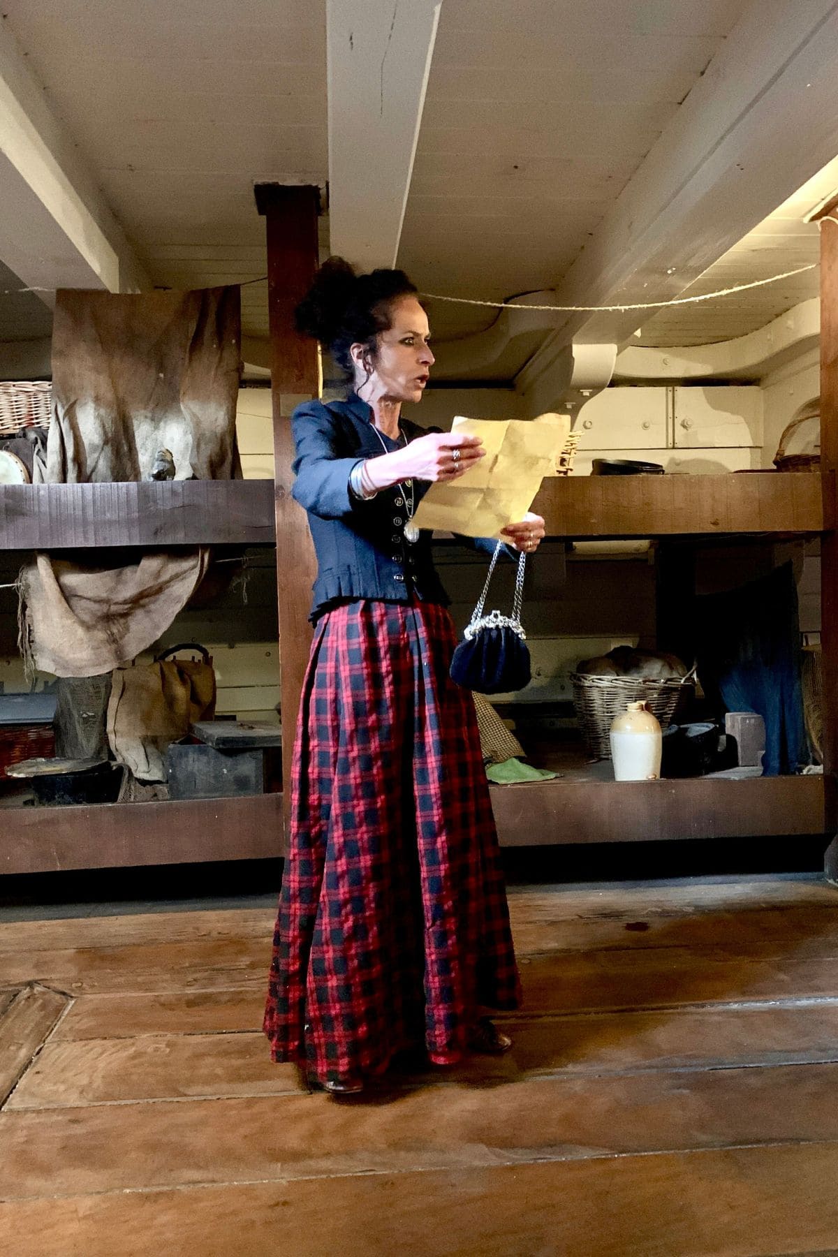 Woman in period costume in the lower part of a ship, reading from a piece of paper.