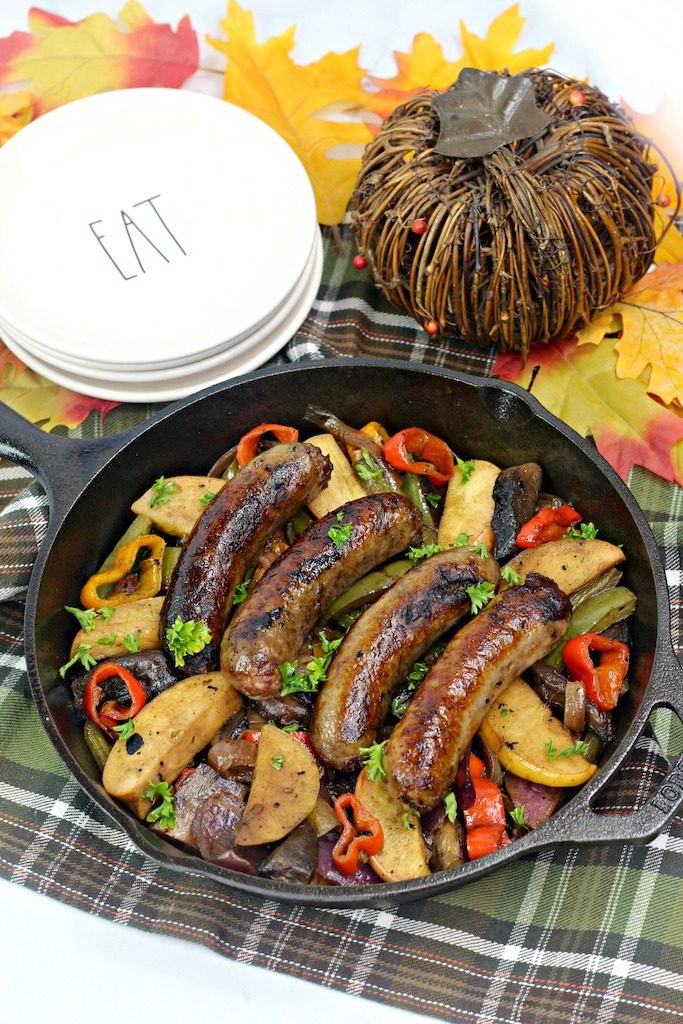 Bratwurst in skillet with apples, peppers, and onions on a plaid cloth with a straw pumpkin and white plates.
