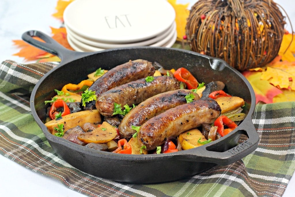 Bratwurst in skillet with apples, peppers, and onions on a plaid cloth.