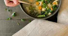 Chicken and vegetable soup in a bowl on a black table with a hand holding spoon.