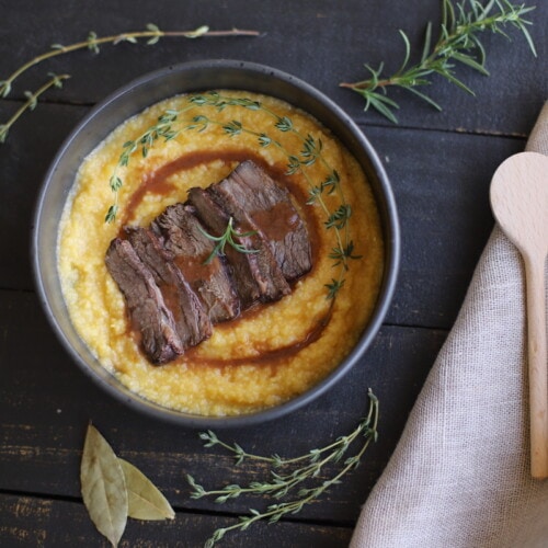 Braised beef over polenta with rosemary, thyme, and juices in a black bowl on a black board.