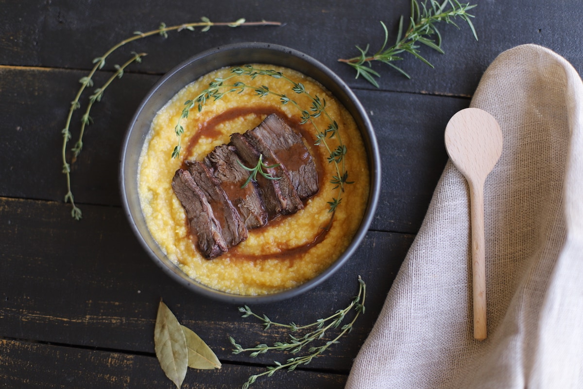 Braised beef over polenta with rosemary, thyme, and juices in a black bowl on a black board.