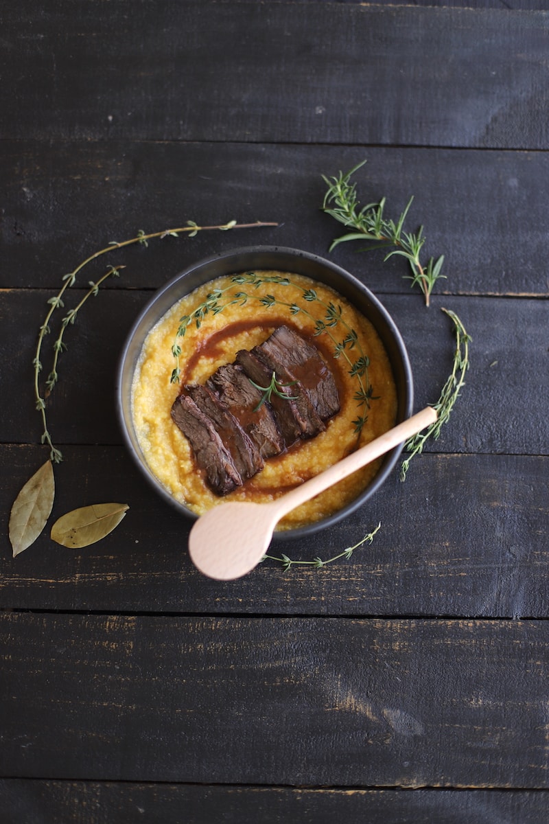 Braised beef over polenta with rosemary, thyme, bay leaves, and juices in a black bowl on a black board.