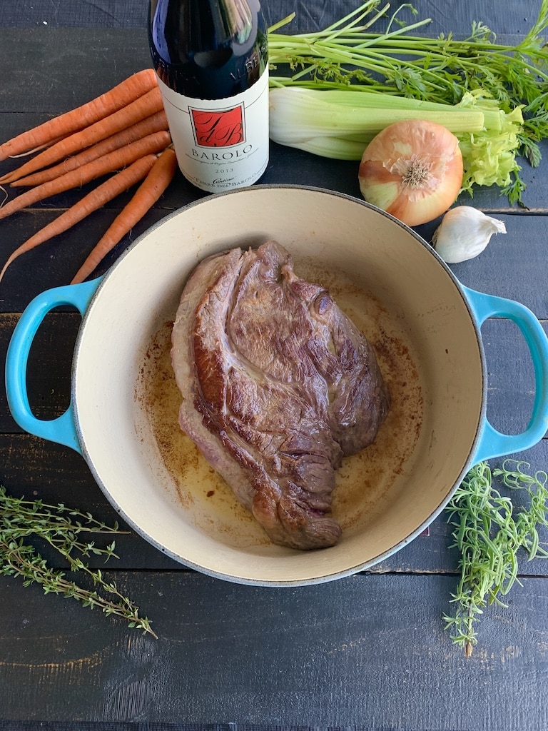 Seared beef roast with raw carrots, onion, garlic, celery, and herbs in a blue pot on a black board.