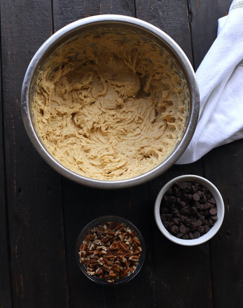 cookie batter with bowl of chocolate chips and bowl of pecans on wood board