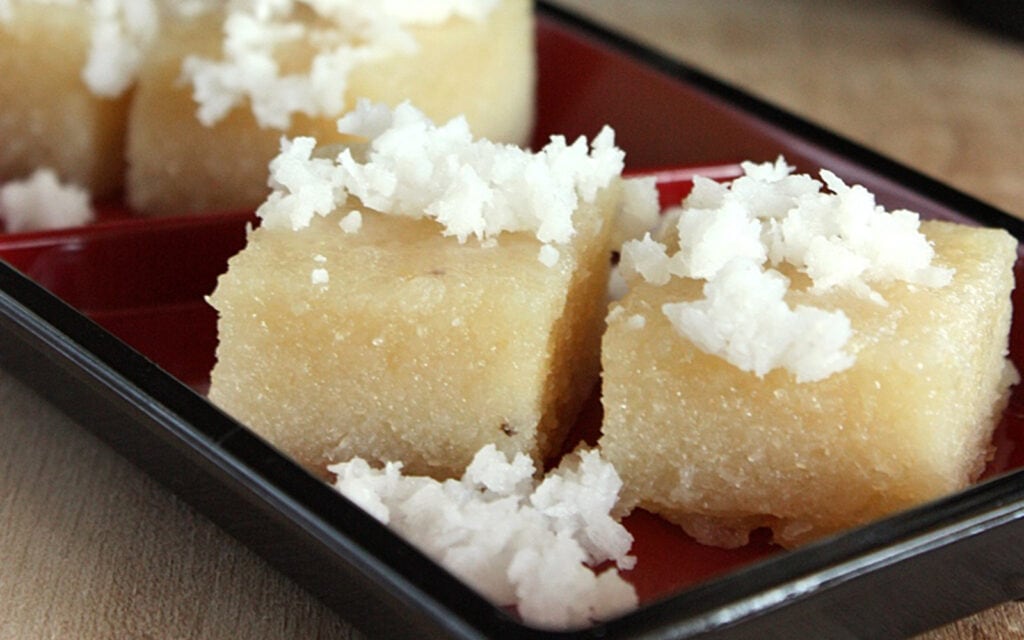 Pieces of cassava cake topped with coconut on a brown tray.