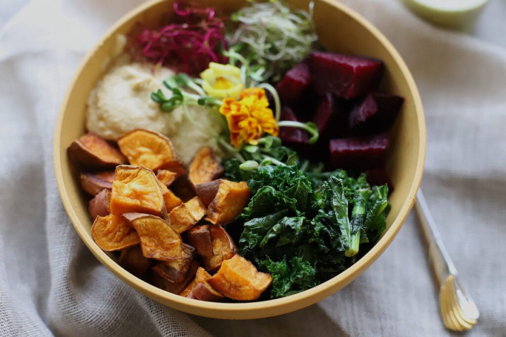 A bowl of mixed vegetables including roasted sweet potatoes, kale, beets, microgreens, and hummus, garnished with an edible flower, placed next to a fork and a small bottle on a cloth surface.