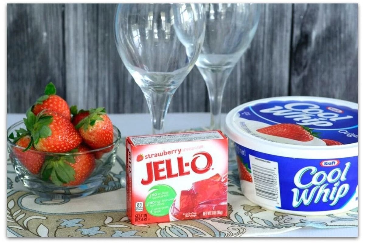 Glass bowl of strawberries, box of jello, Cool Whip topping, and two tall glasses on a table.