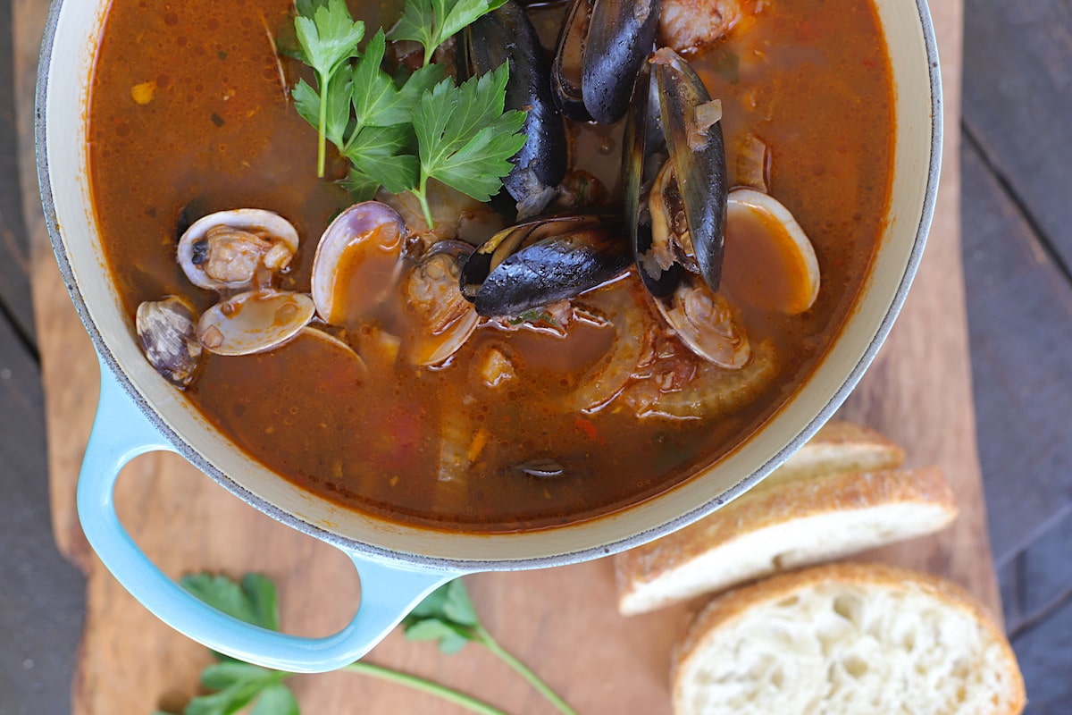 Blue pot of tomato based seafood stew sitting on a wood board with bread.