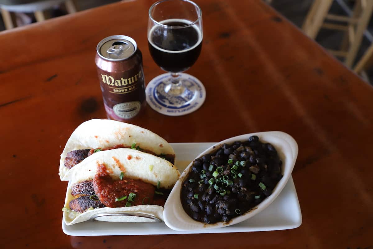 Fish tacos with black beans on side and a beer can and glass on wood table.