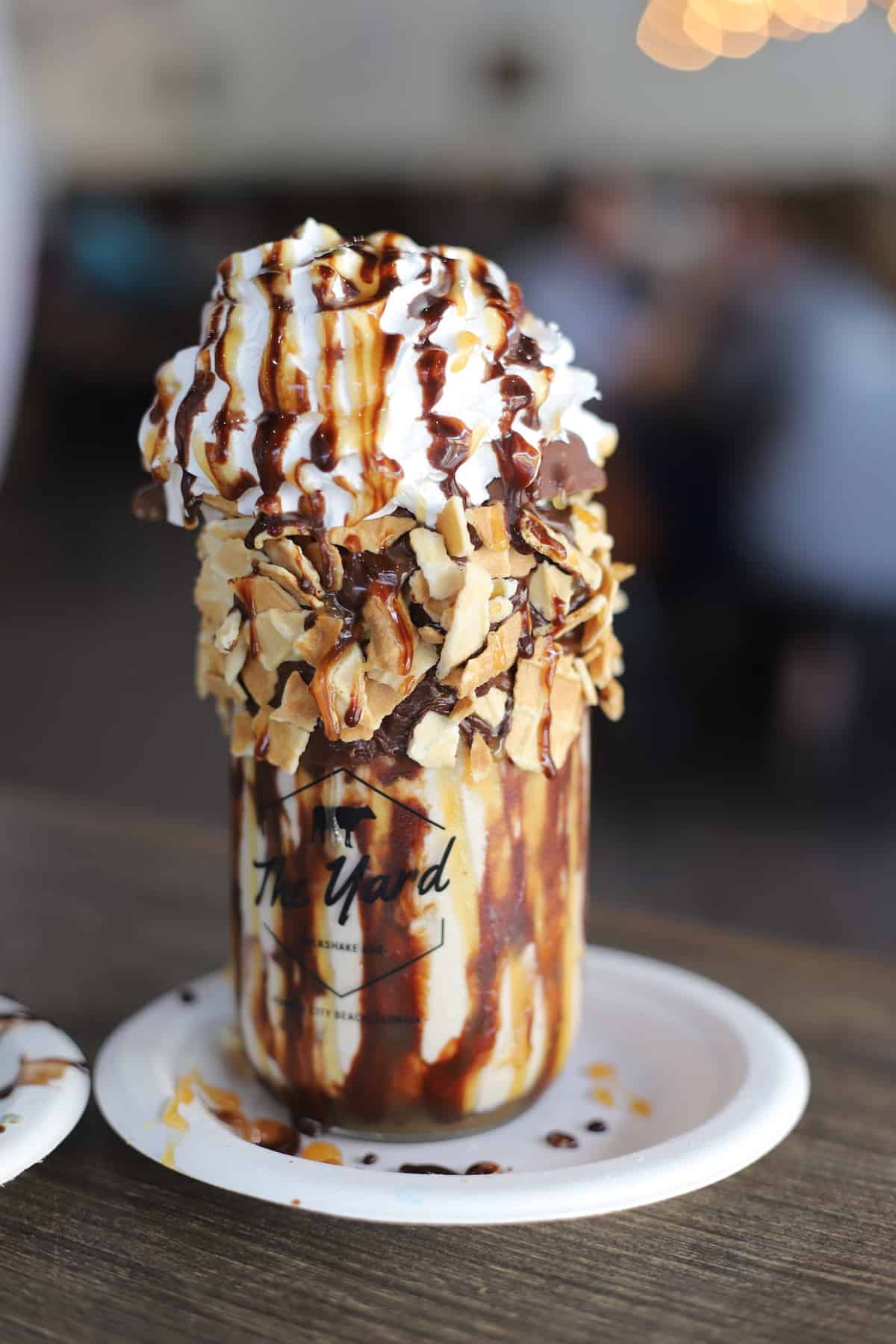 Milkshake in a mason jar with whipped cream,, chocolate sauce, and nuts on a white plate.
