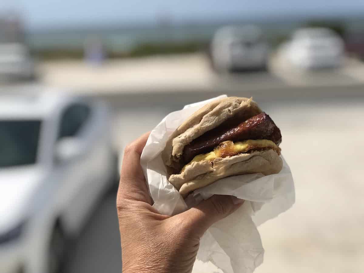 English muffin with egg and sausage and beach in background.