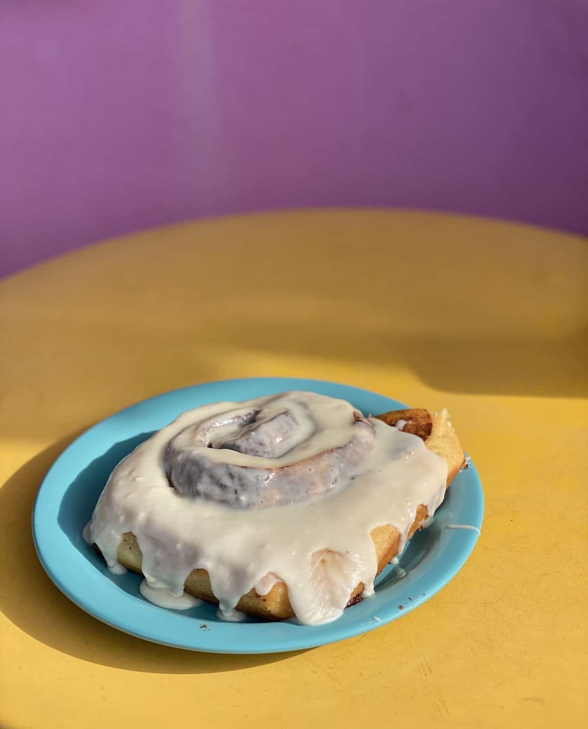 Large cinnamon bun on yellow table with purple background.