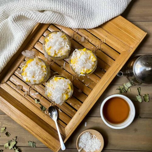 Mango with coconut in a jar topped with crushed ice on a bamboo board with a white cloth and other ingredients.