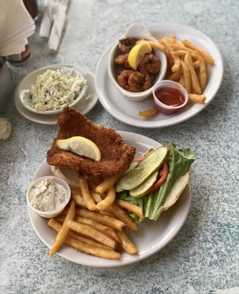 Grouper fish sandwich and fries and shrimp platter with fries and coleslaw on white plates on grey speckled table.