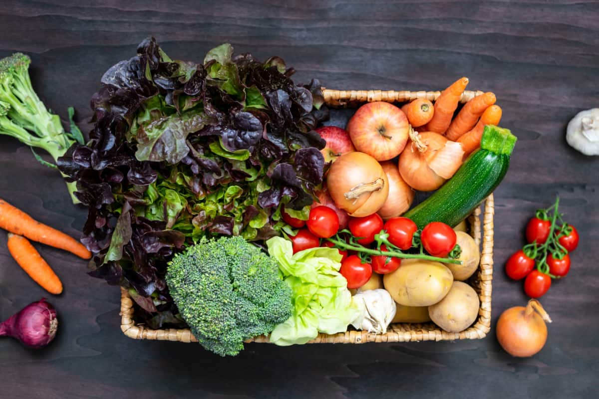 Rectangular wicker basket full of vegetables with cherry tomatoes, onion, carrots, garlic, and red onion on table.