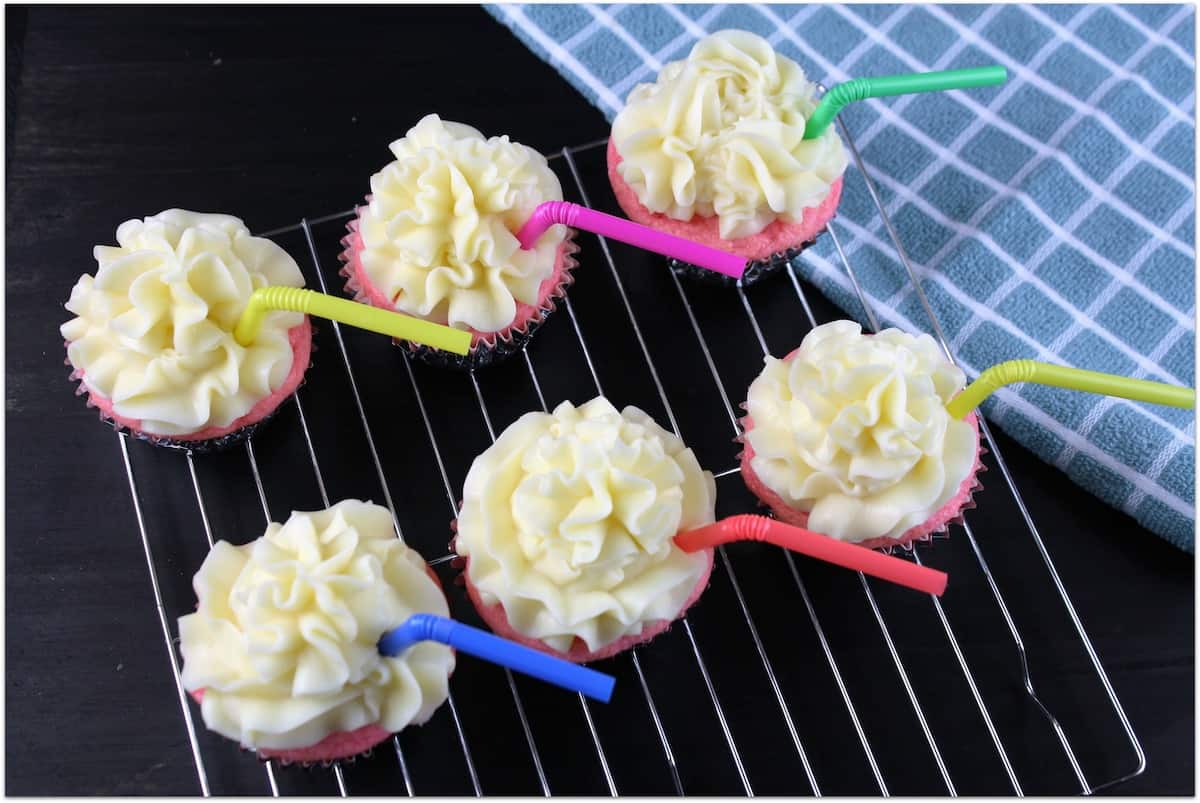 Pink cupcakes with yellow frosting with multi-colored straws in each one, set on a wire rack.