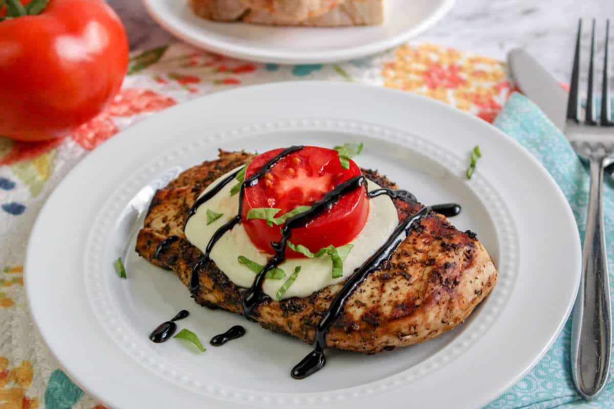 Grilled chicken with tomato and mozzarella, basil, and balsamic glaze on top, on white plate with tomatoes and grilled bread.