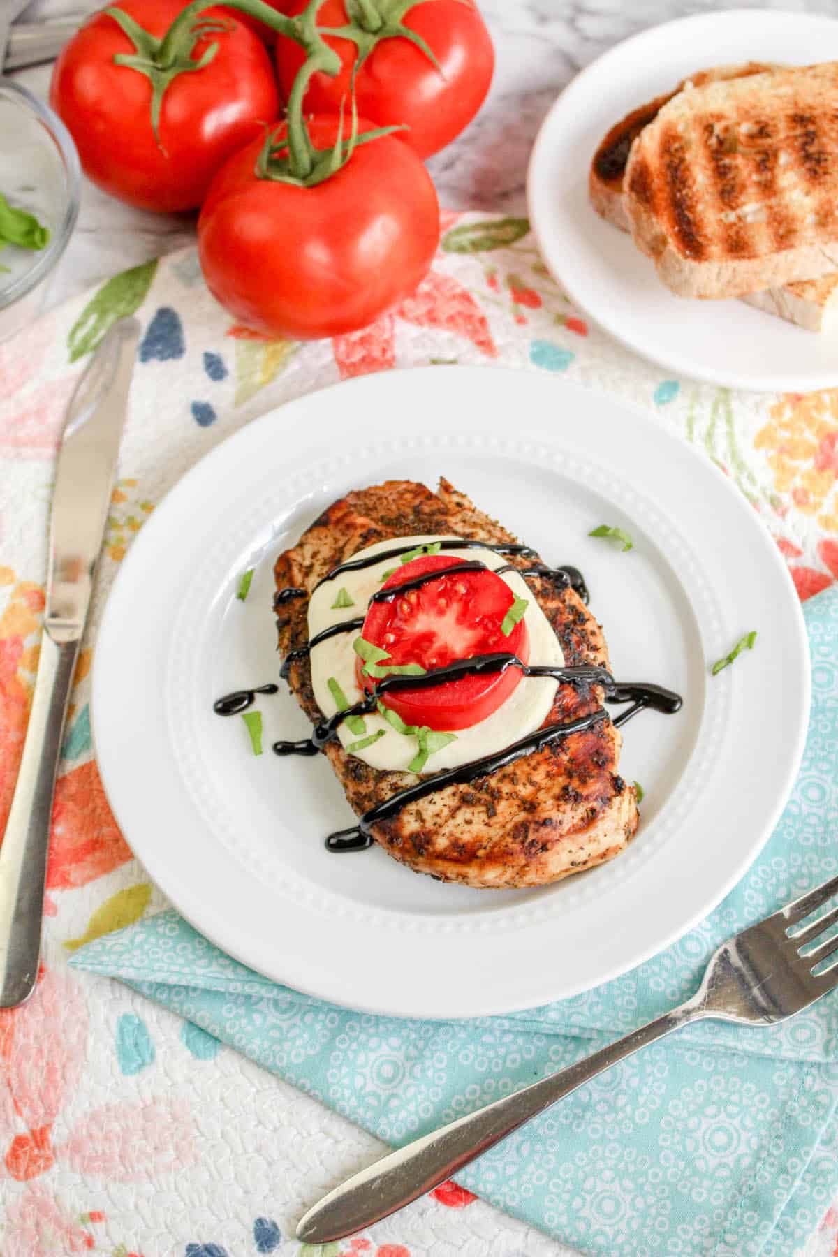 Grilled chicken with tomato and mozzarella, basil, and balsamic glaze on top, on white plate with tomatoes and grilled bread.