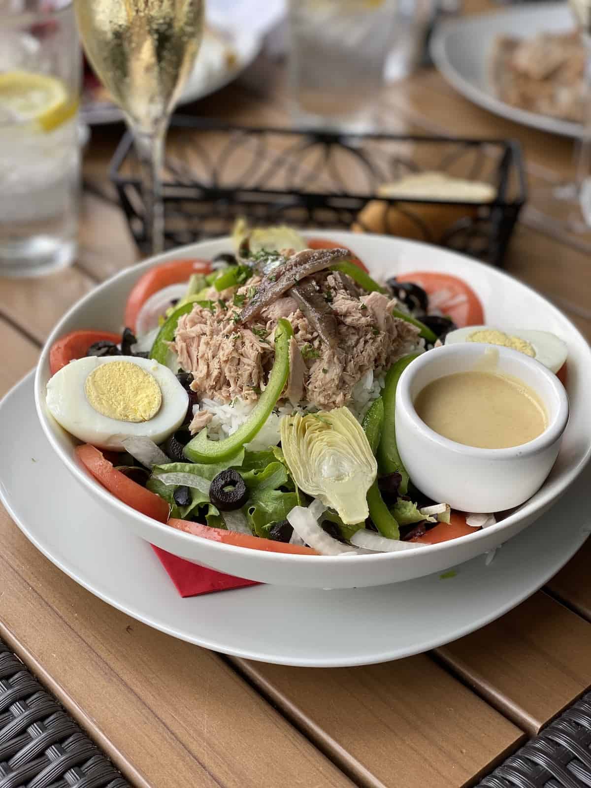 Salad with tuna, egg, artichokes, olives, tomatoes, with a small bowl of dressing on the side.