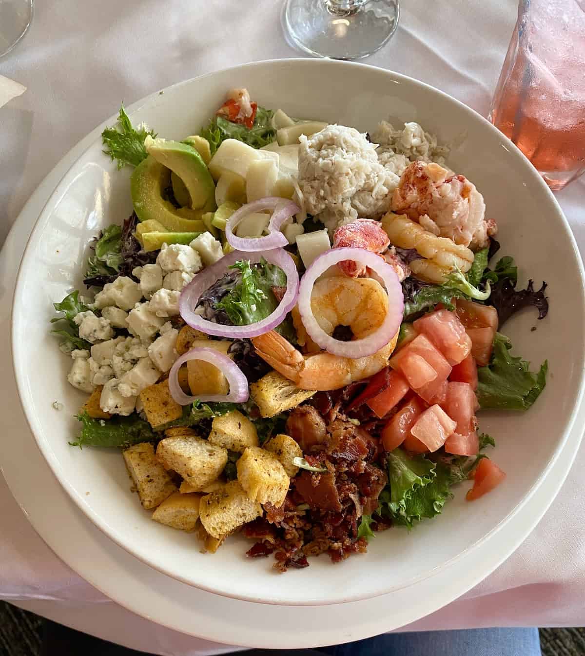 Salad with bacon, cheese, tomatoes, shrimp, crab, lobster, and avocado in a white bowl.