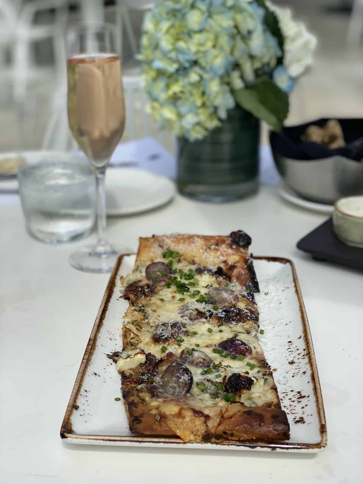 Flatbread with cheese and grapes on white stone platter with glass of Prosecco.