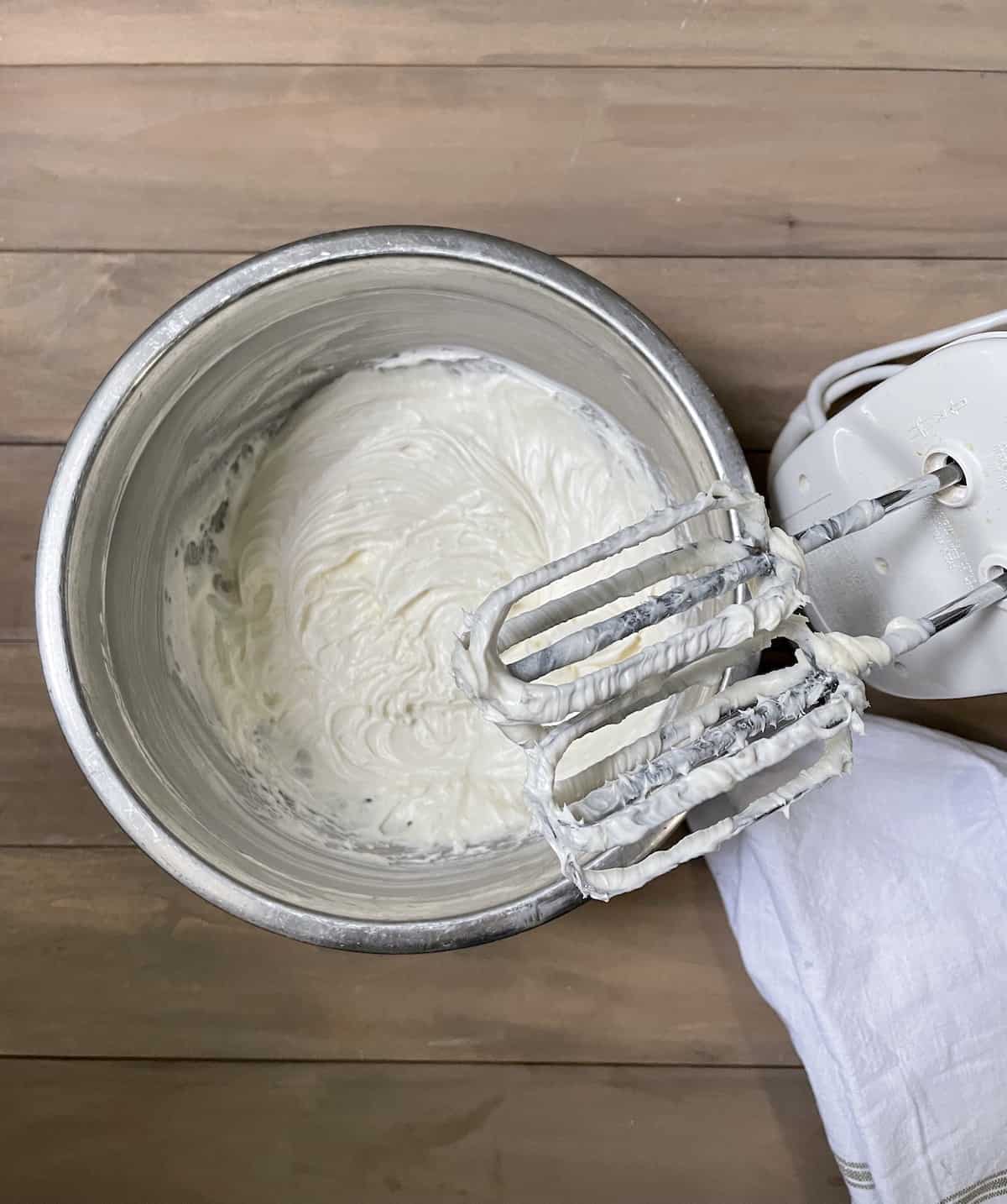Cream cheese frosting in stainless bowl with electric hand mixer.