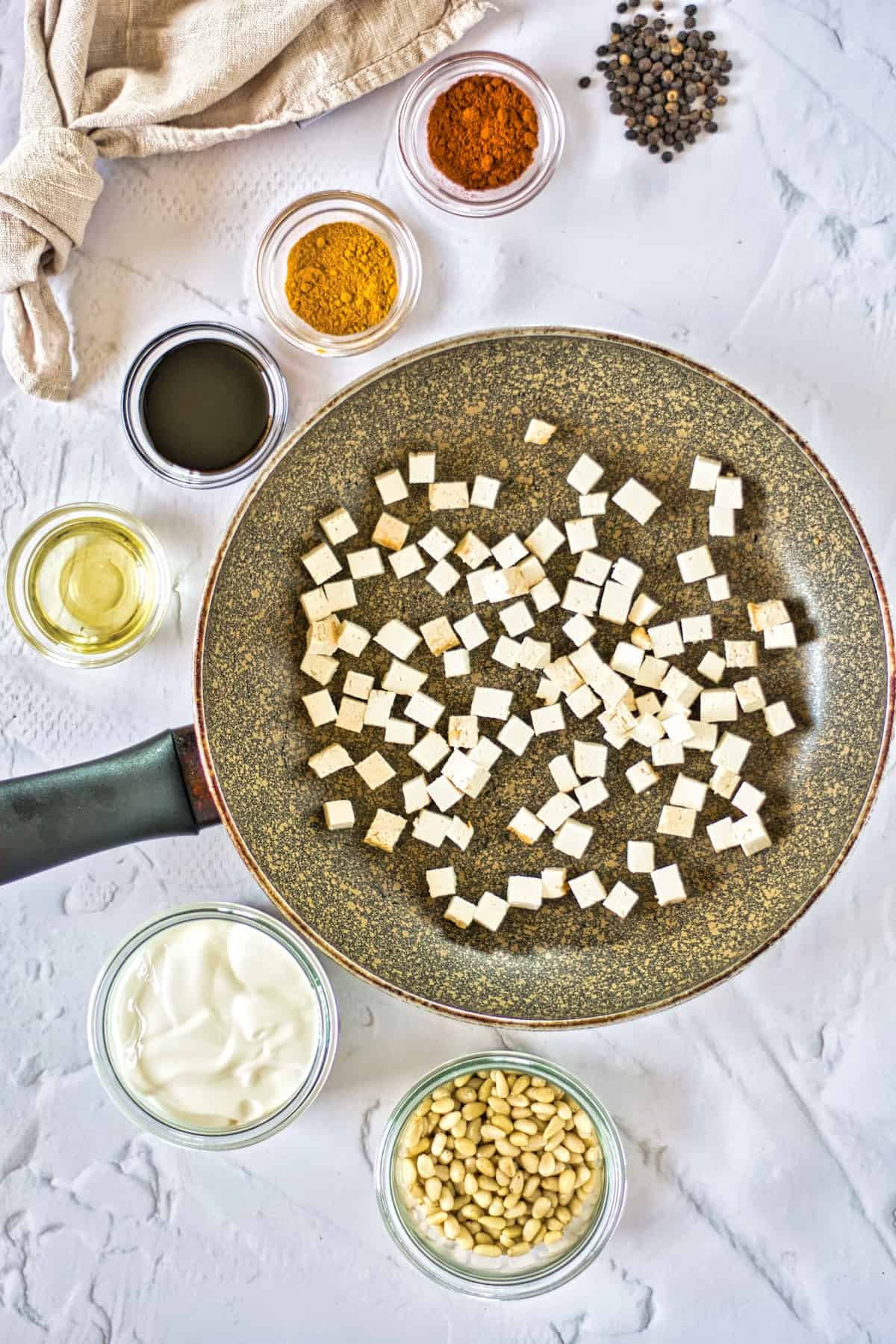 Napkin, seasoning, peppercorns, oil, tofu in pan, vegan cream, and pine nuts, on a white table.