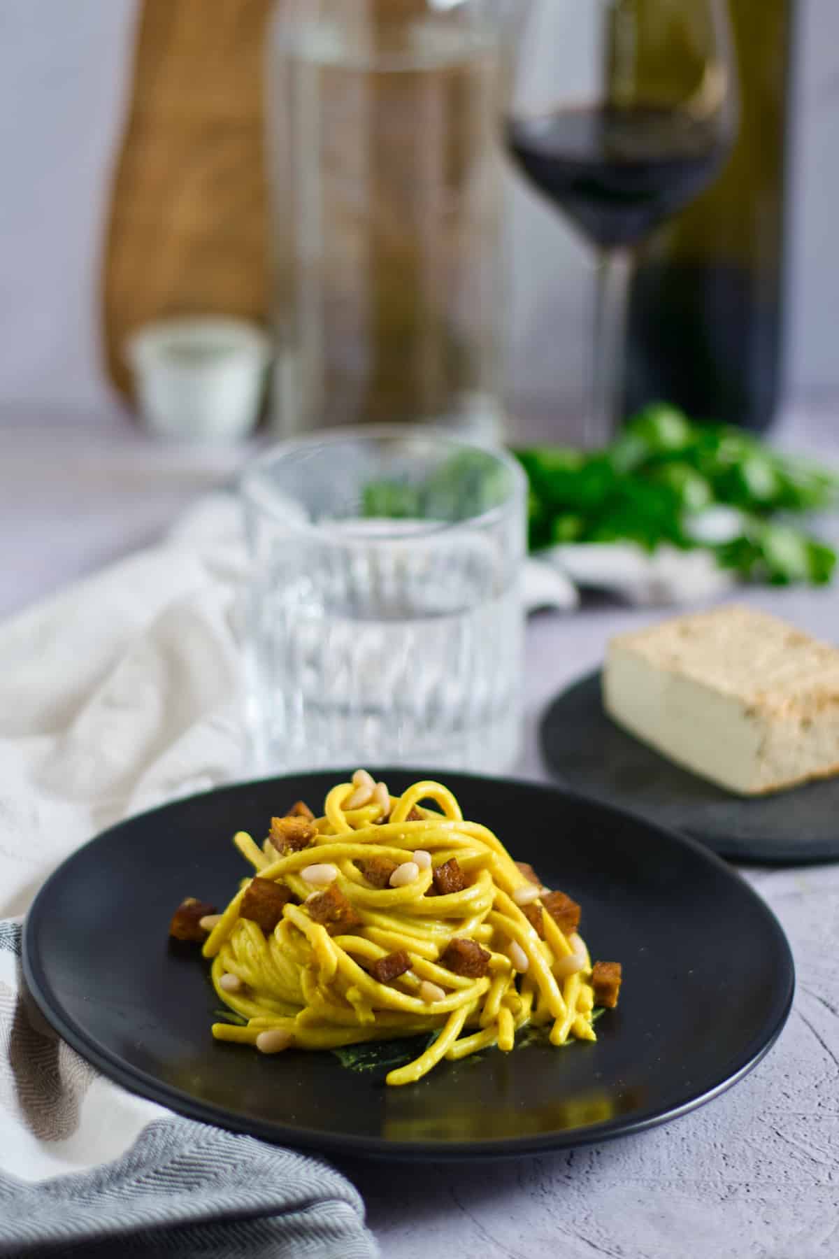 Pasta with tofu and pine nuts on a black plate with glass of water in background.