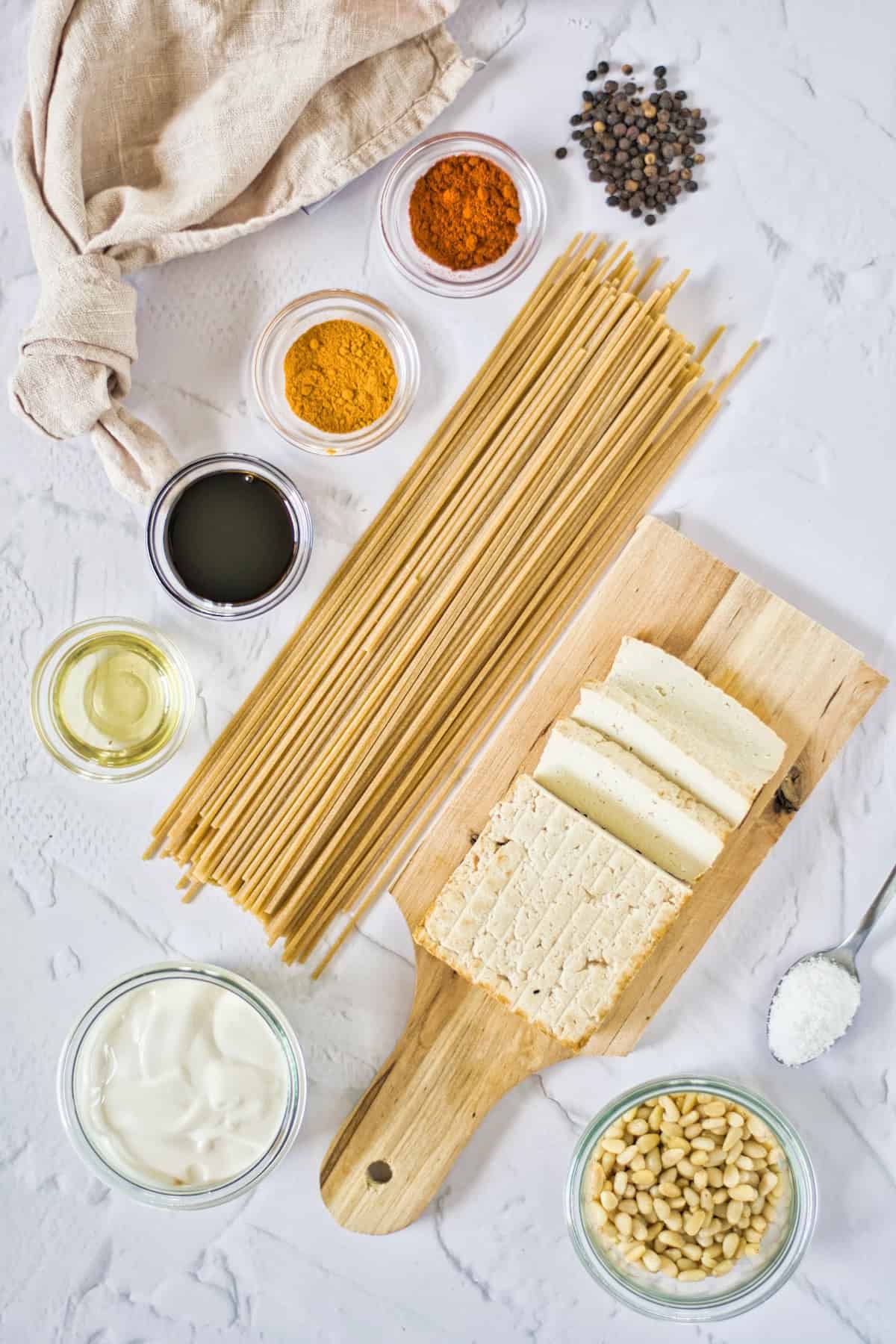 Dried pasta, seasoning, tofu, peppercorns, vegan cream, pine nuts, salt on a white table.