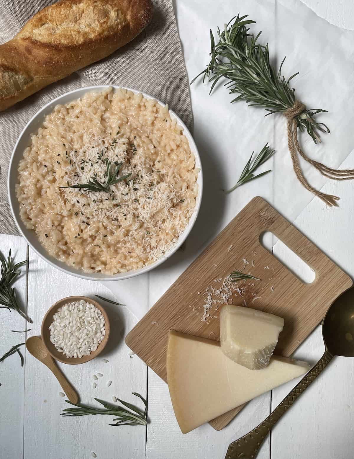 Risotto with cheese in a white bowl with rice, cheese on cutting board, rosemary, bread, on a white table.