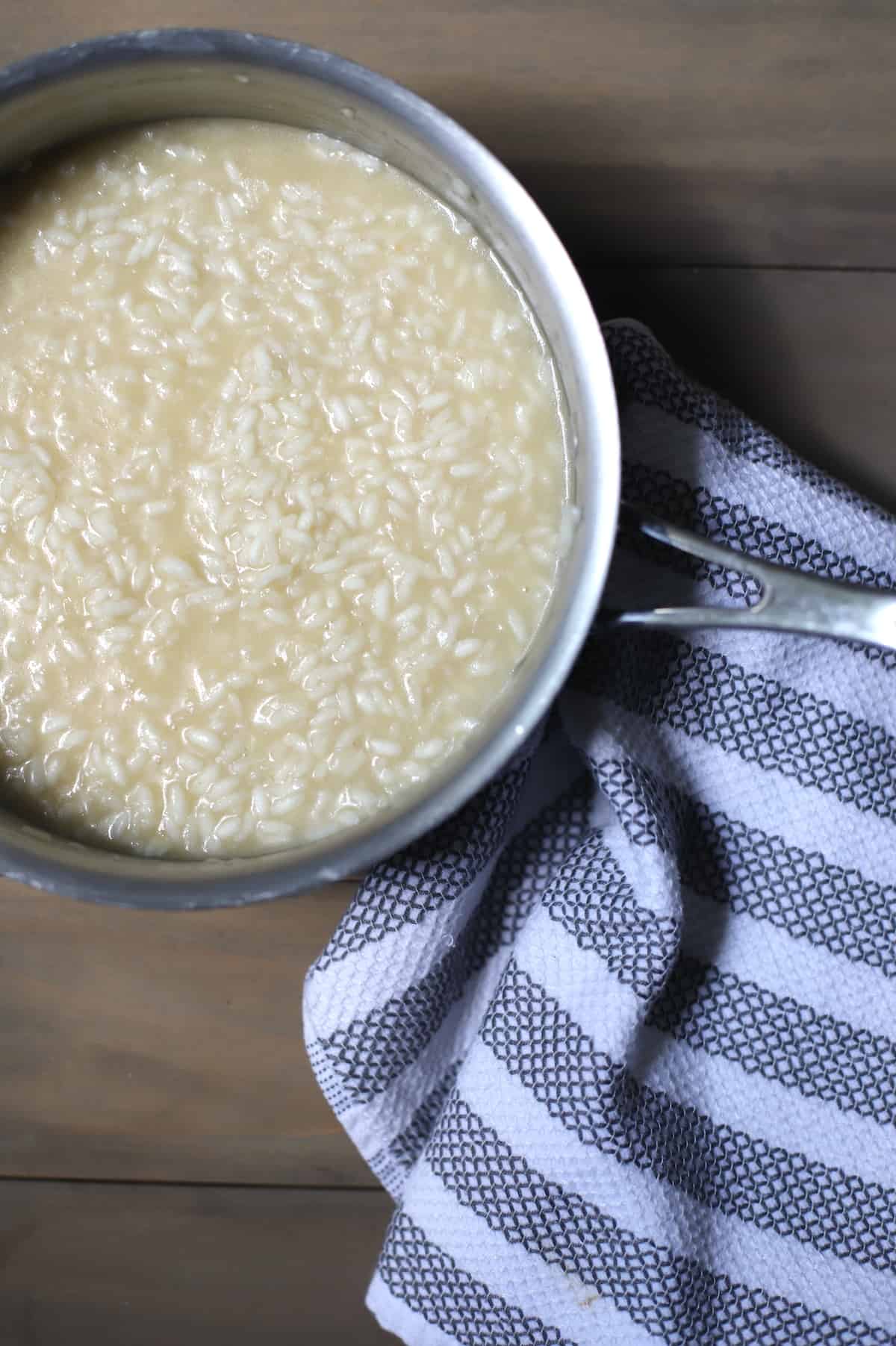 Arborio rice in a pot with broth on a wood board with a white and blue tea towel.