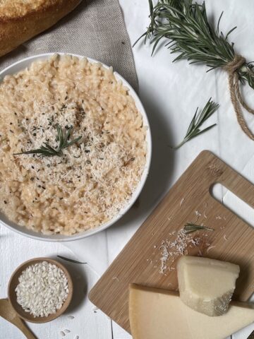 Parmesan risotto in a white bowl with rosemary, cheese on a wood board, and rice, on a white table.