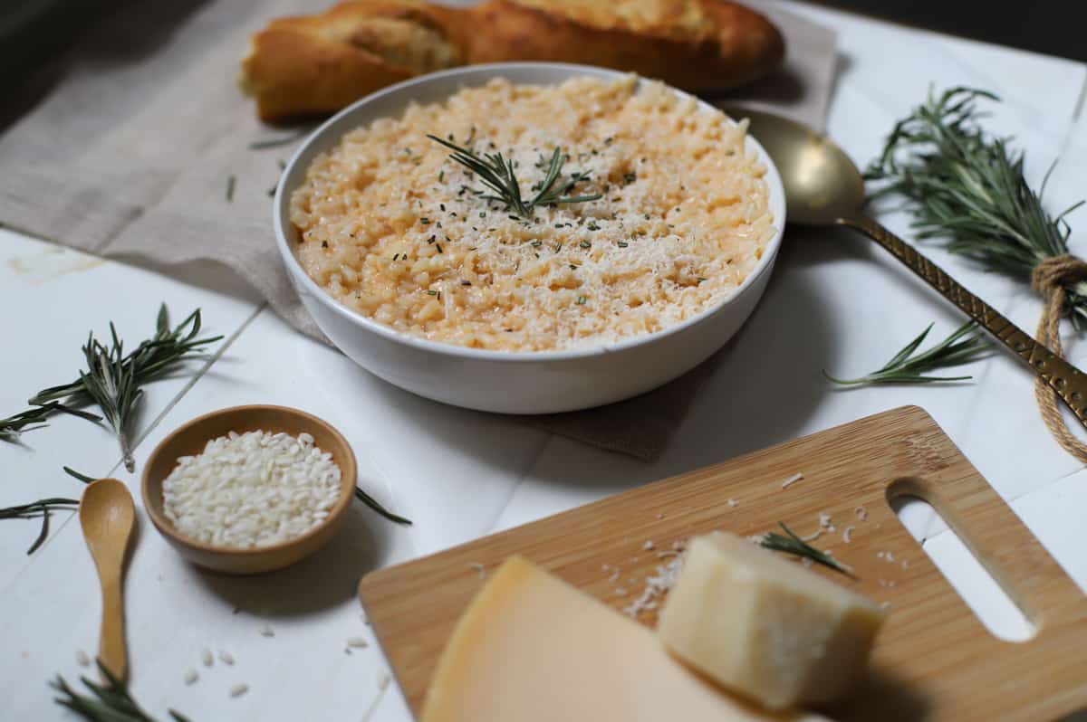 Risotto with cheese in a white bowl with rice, cheese on cutting board, rosemary, bread, on a white table.