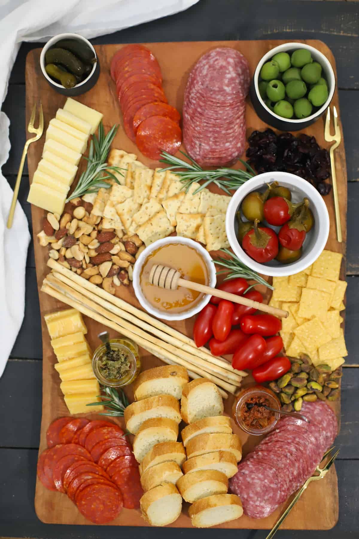 Charcuterie board with salame, pepperoni, cheeses, spreads, nuts, pickles, olives, and rosemary.