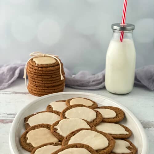Iced cookies on a white plate with a stack of cookies and a bottle of milk in background.